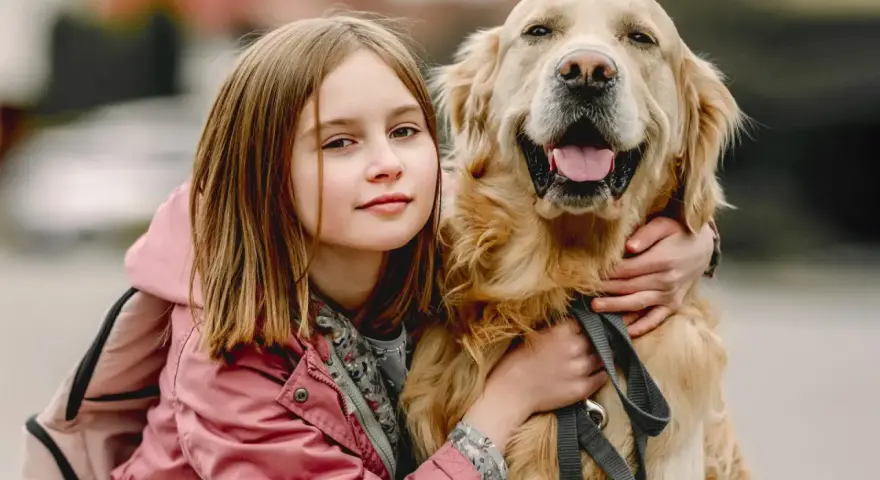 Dziewczynka w różowej kurtce przytula uśmiechniętego golden retrievera.